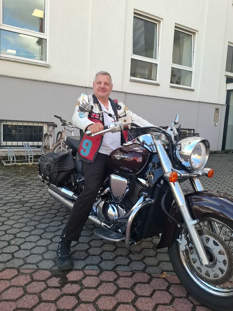 Herr Dähne sitzt auf einem dunkelroten Motorrad vor einem Gebäude und blickt zur Kamera.